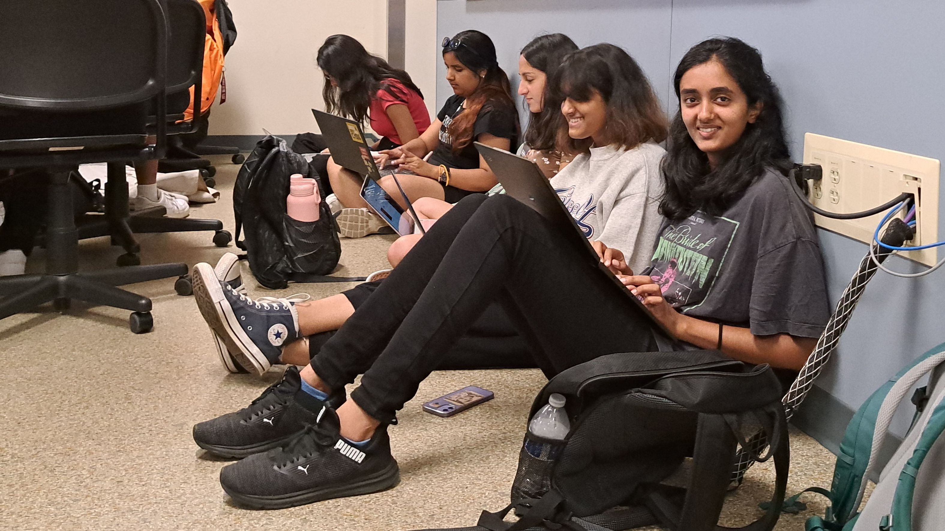 A group of 5 people sitting on the floor with laptops in their laps at a workshop. The person closest to the camera is looking at the camera and smiling